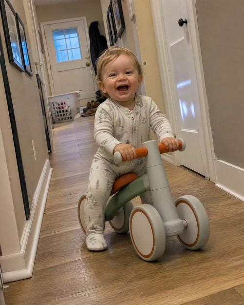 Laughing one-year-old in pajamas riding the Montessori balance bike down the hallway at home